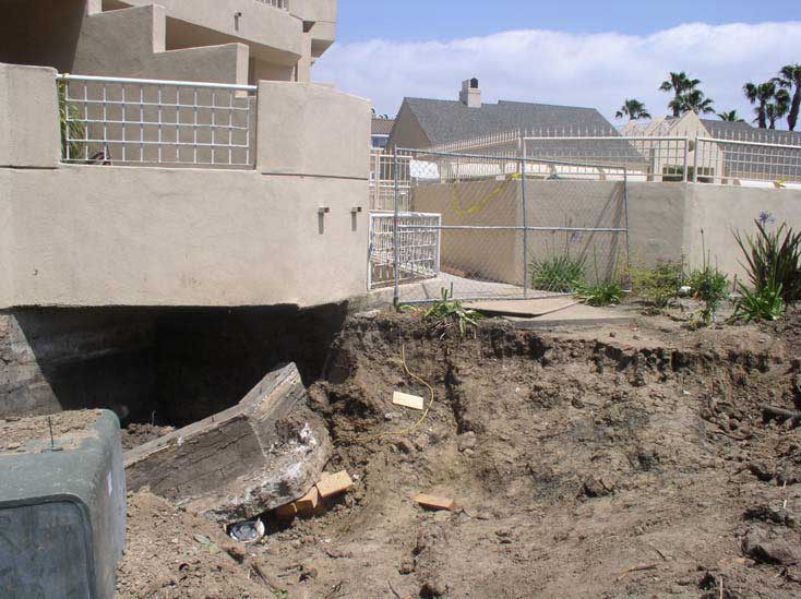 A chain link fence blocks the sidewalk where the earth has given way into a large, deep pit that extends down several feet from the ground level of an apartment building.