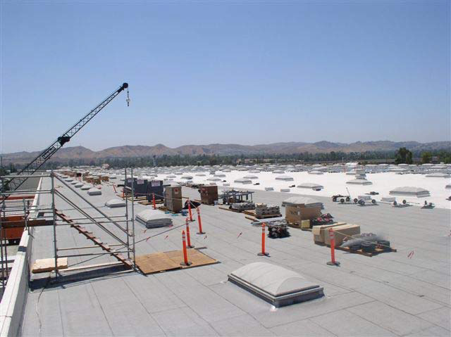 A large, flat, white building rooftop is covered with dozens of skylights. A crane hovers over a section of the roof.