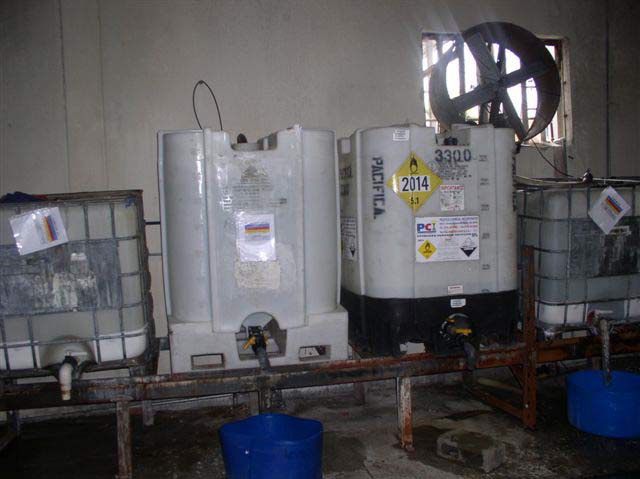 Four large, white plastic tubs sit on a low, rusted metal rack in a room with a large ventilated circular opening in the wall.