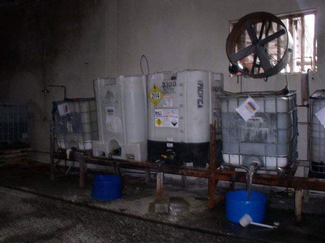 Four large, white plastic tubs sit on a low, rusted metal rack. Two of the tubs have blue plastic buckets underneath spout openings at the bottom of the tubs.