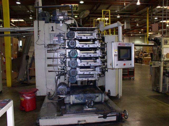 A large printing machine in a warehouse with the six ink trays exposed. There are large round knobs on the left of the ink trays.