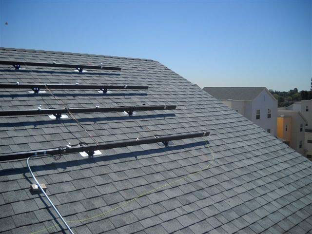 Four horizontal mounting brackets for solar panels are attached to the steeply slanted roof of a building, and there are other multi-story residential buildings in the distance.