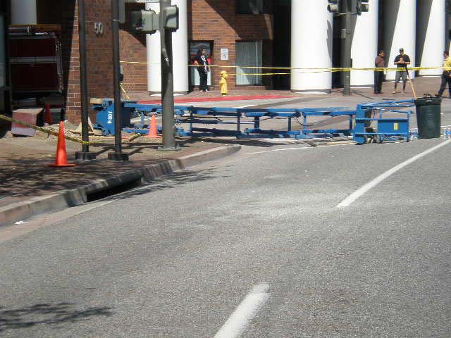 An extended blue lift lies across a sidewalk and a street next to an upright black garbage pail.