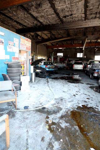 Entrance of automotive shop where incident occurred, showing seven vehicles parked at angles, tires piled up, two chairs facing outward, and a long hose running to the back of the space.