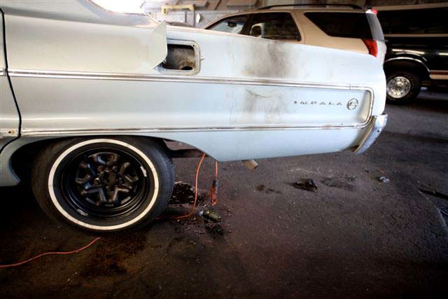 Left rear quarter of vehicle is shown with fuel filler door open, and behind the rear wheel is an orange electrical cord draped over the exhaust pipe with the exploded drop light hanging down and touching the ground.