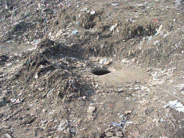 An opening to a manhole leading to the draining system is surrounded by dirt and debris, with an area of exposed concrete where debris has been swept away from the opening.