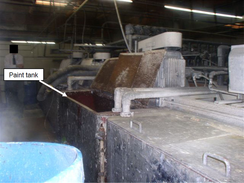 A paint-splattered metal tank with three open metal panel lids sits in a factory building.