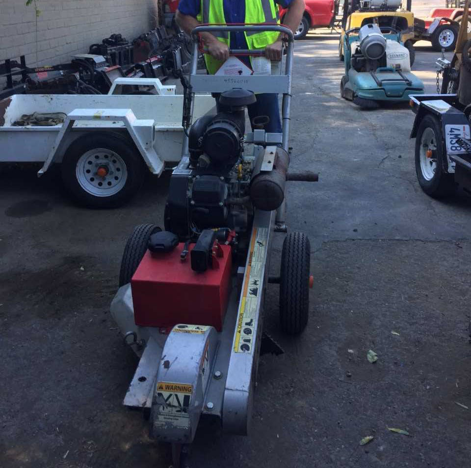A metal and red machine on two wheels - the size of a large lawnmower - is pushed by a worker. There are multiple warning signs along one side of the machine.