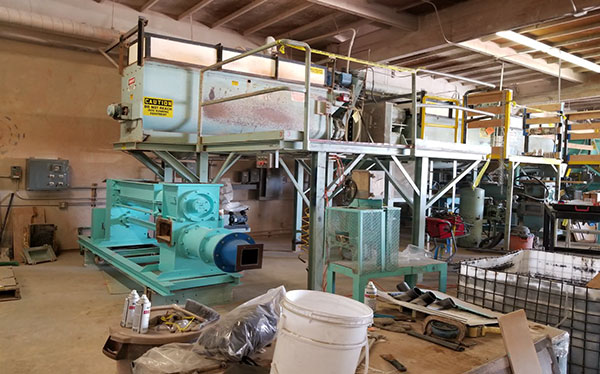A large machine stands in a warehouse with buckets and industrial parts stacked in front of it.