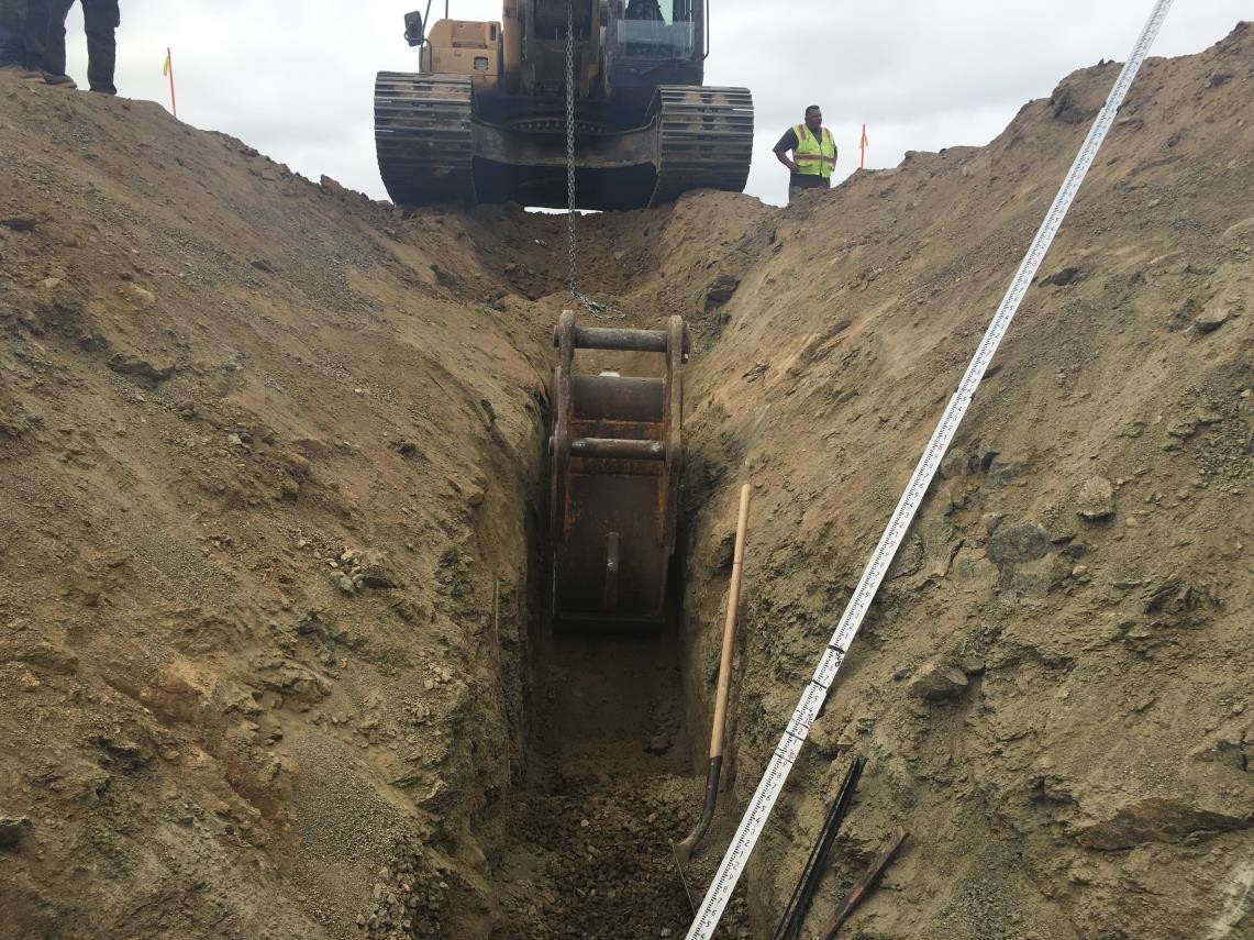 view of trench from above with excavator nearby and metal rod indicating its slope and depth